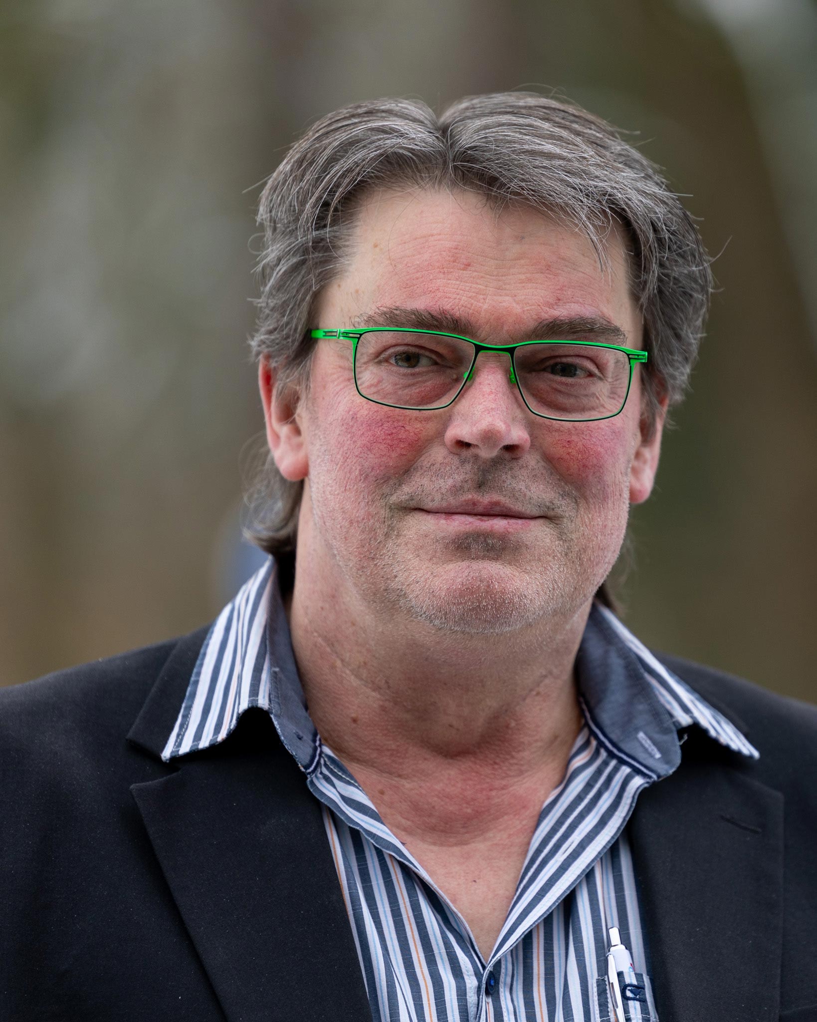 Ein grauhaariger Mann mit grünem Brillengestell und leichtem Lächeln trägt ein gestreiftes Hemd und einen dunklen Blazer