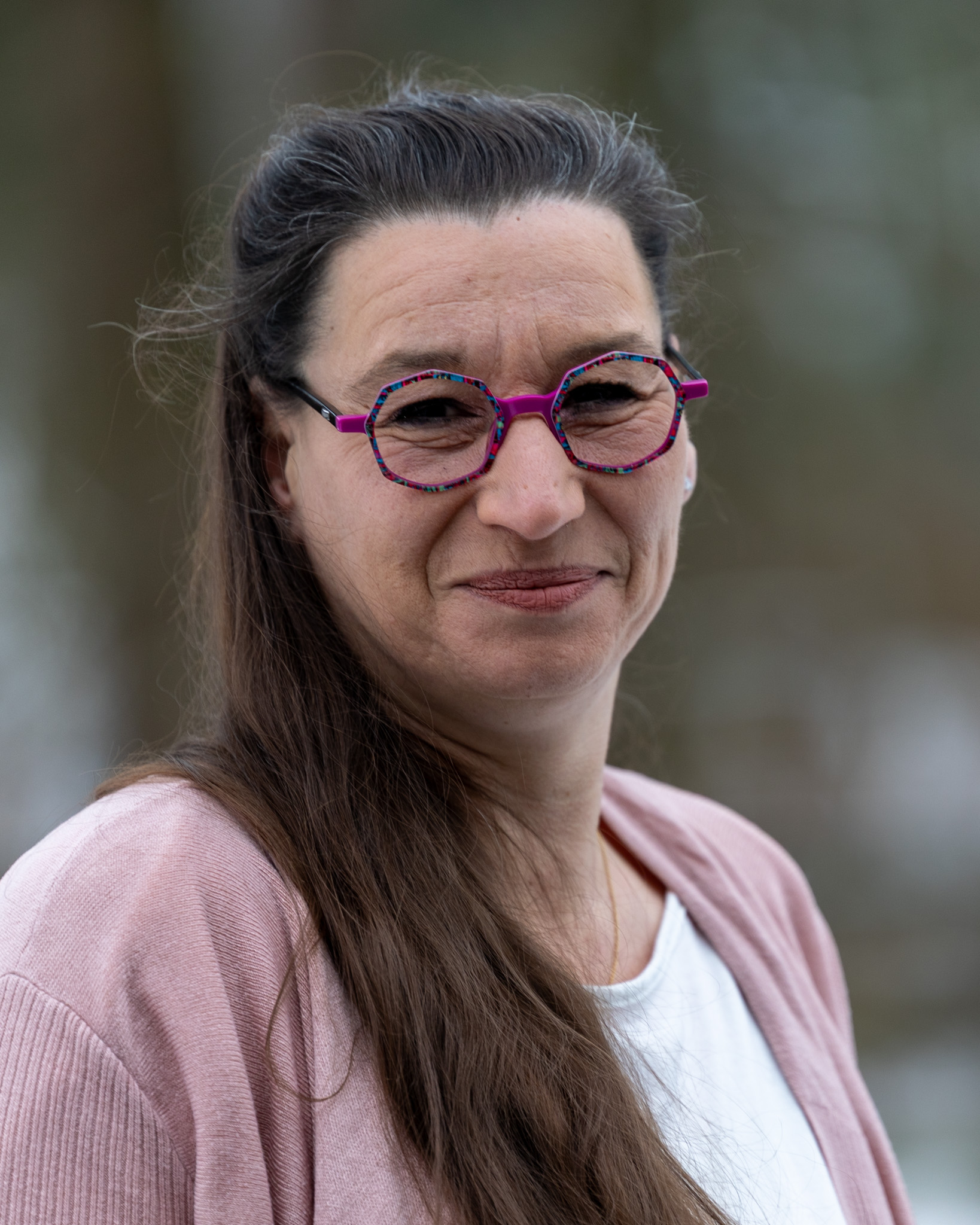 Eine Frau mit langen braunen Haaren und auffälliger rosa Brille lächelt freundlich